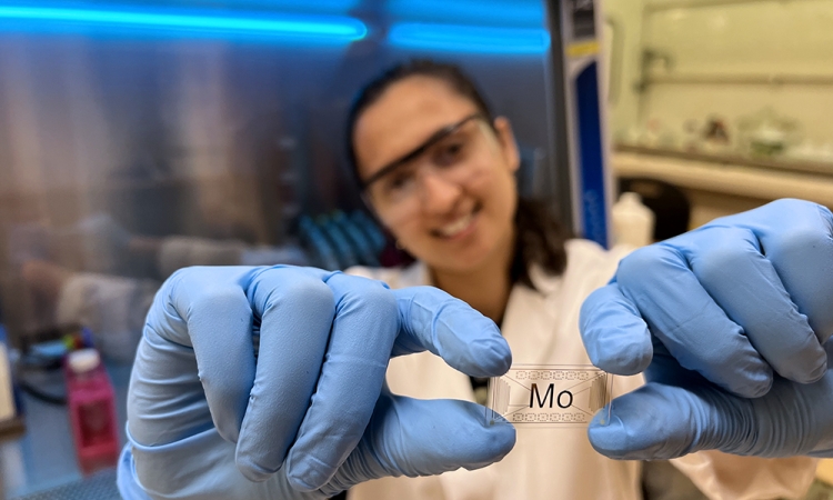 A woman wearing protective glasses and latex gloves holds a glass chip that says "Mo"