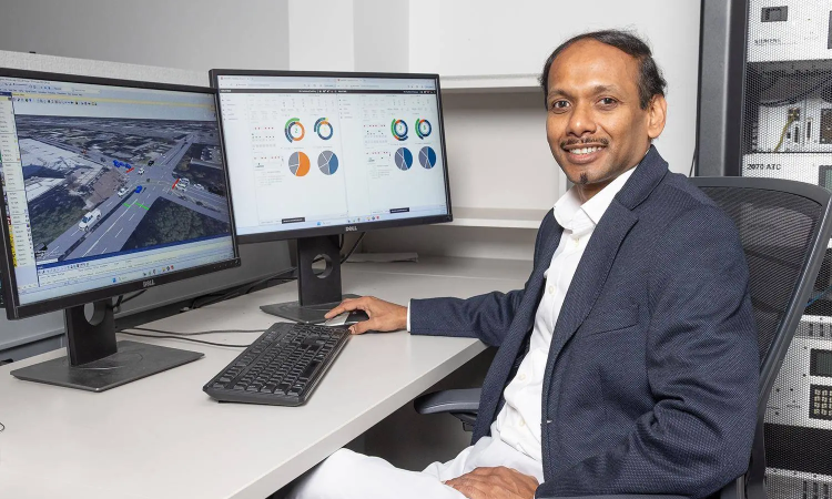 A man sits next to two computer monitors showing pie graphs and a road intersection