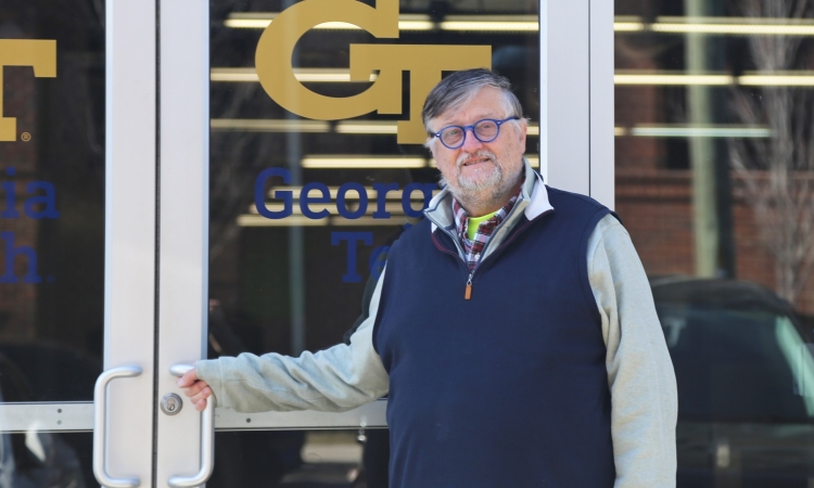 A man holds a door handle with a gold interlocking GT logo on the glass behind him