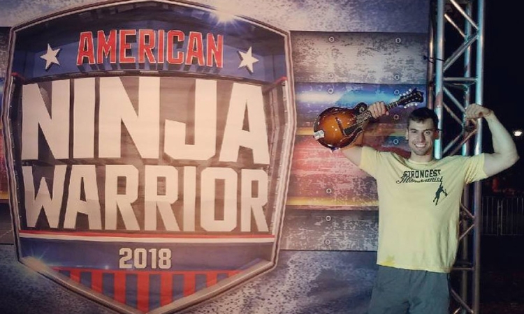 Civil engineering undergrad Elie Cohen flexes his biceps while holding his mandolin in front of an American Ninja Warrior 2018 sign. (Photo Courtesy: NBC)