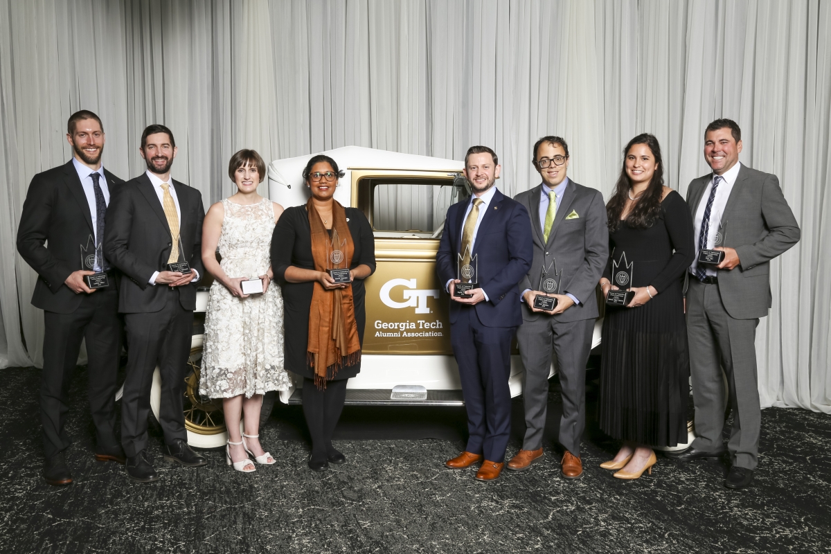 A group of people poses in front of an antique gold car and a white curtain