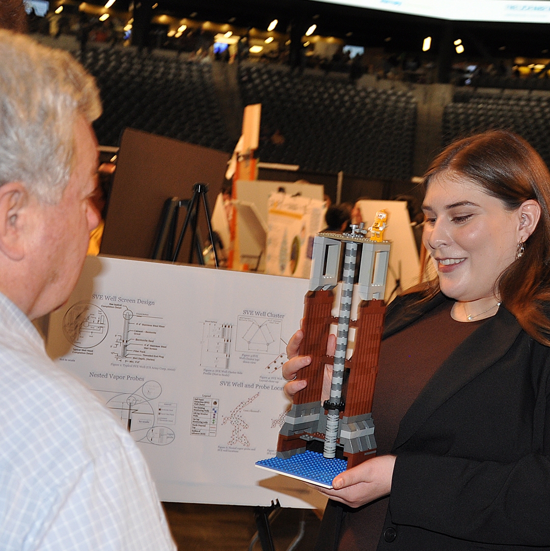 Rebecca Chapman, wearing a black blazer and a maroon top, shows a Lego model of the project to a Capstone visitor.